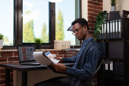 african-american-startup-owner-with-glasses-holding-clipboard-paper-with-bussiness-charts-comparing-data-with-sales-results-laptop-entrepreneur-analyzing-marketing-indicators-laptop